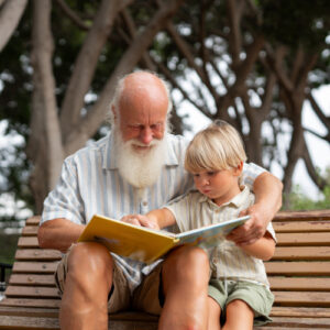 An elderly man with a white beard reads a picture book to a young boy on a wooden bench outdoors, surrounded by trees.
